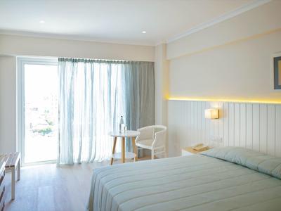 Lichte hotelkamer met tweepersoonsbed, stoel, tafel en toegang tot een balkon met gordijnen.