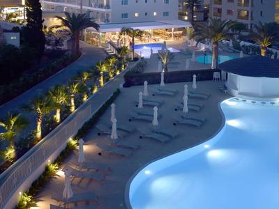 Verlichte hotelzwembadgebied 's nachts met ligstoelen en omliggende gebouwen.