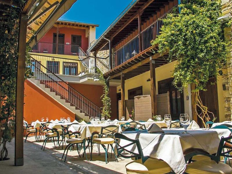 Innenhof mit gedeckten Tischen, Stühlen, Treppe, Balkonen und bunten Häusern bei klarem Himmel.