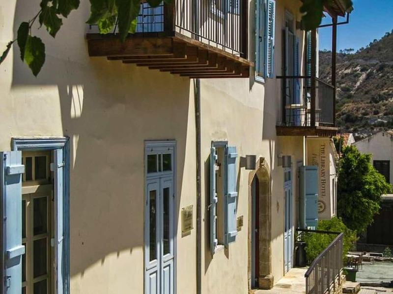 Haus mit blauen Fenstern und Türen, Balkon und Sonnenlicht, umgeben von Pflanzen.