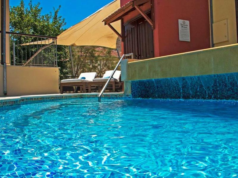 Klares blaues Wasser in einem Pool mit Treppe, Sonnenschirm und Liegen im Hintergrund vor Hauswand.