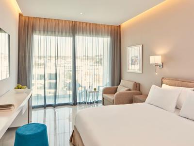 Modernes Schlafzimmer mit großem Bett, beiger Wand, blauem Hocker und bodentiefen Fenstern.