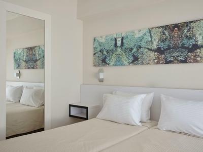 Hell eingerichtetes Schlafzimmer mit weißem Bett, Kissen, modernem Wandbild und großem Spiegel.