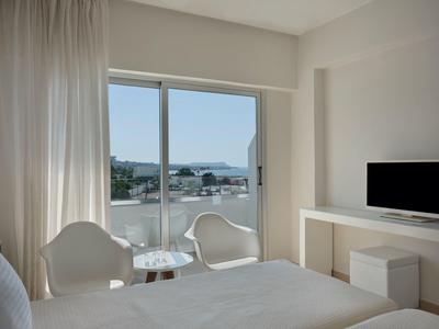 Helles Schlafzimmer mit Doppelbett, zwei weißen Sesseln, Tisch und Fernseher vor Balkon mit Meersicht.