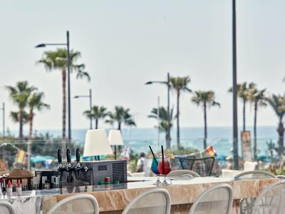Terrasse eines Hotels mit Tischen, Stühlen und Blick auf Palmen und das Meer.
