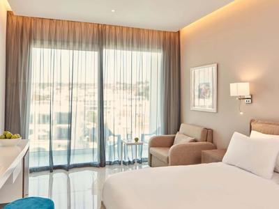 Nowoczesny pokój hotelowy z dużym łóżkiem, fotelami i oknami od podłogi do sufitu.