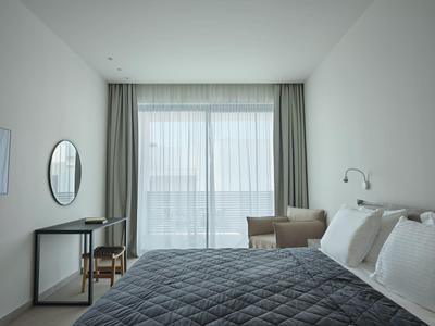 Helles, minimalistisches Schlafzimmer mit grauer Bettdecke, Stuhl, Tisch und großem Fenster mit Vorhängen.