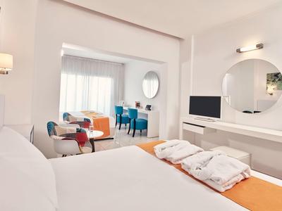 Helles Schlafzimmer mit weißem Bett, orangefarbener Decke, pinken Sesseln und blauem Esstisch.