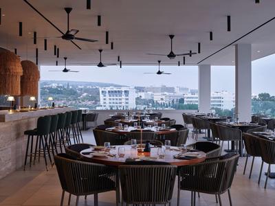 Modernes Restaurant mit Blick auf die Stadt, Holzmöbel und Deckenventilatoren an der offenen Terrasse.