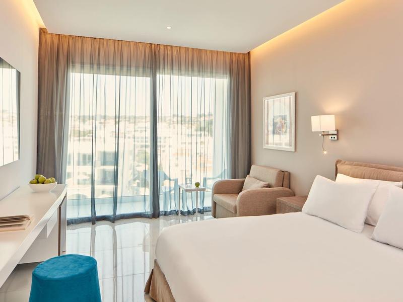 Modernes Schlafzimmer mit großem Bett, beiger Wand, blauem Hocker und bodentiefen Fenstern.