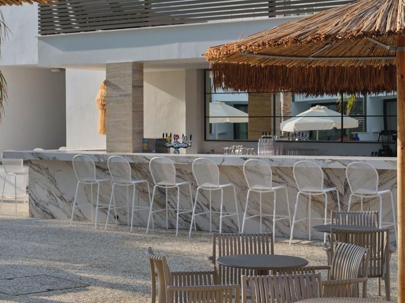 Moderner Außenbarbereich mit hohen weißen Barstühlen und runden Holztischen unter Strohdach.