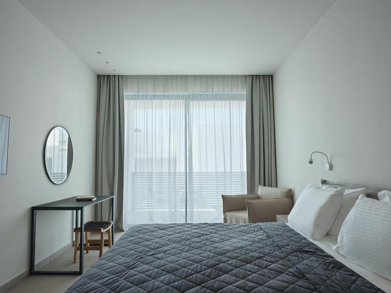 Helles, minimalistisches Schlafzimmer mit grauer Bettdecke, Stuhl, Tisch und großem Fenster mit Vorhängen.