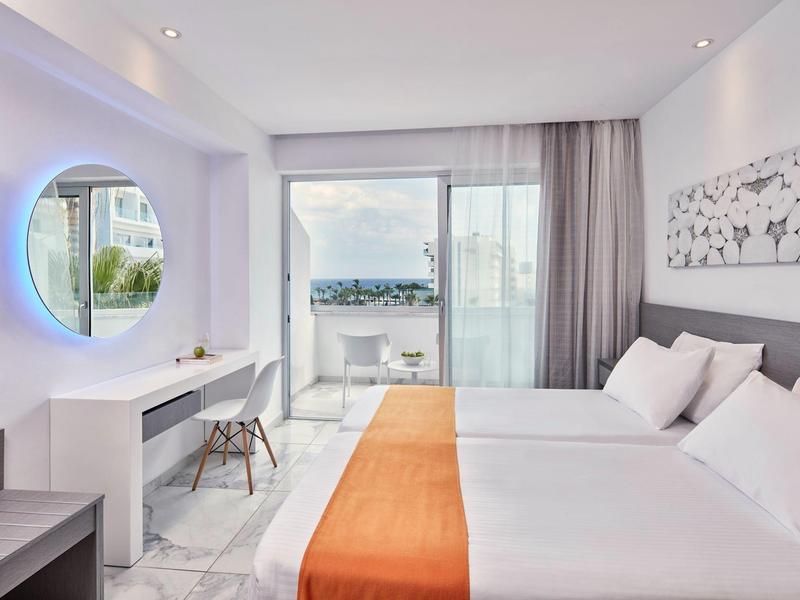 Helles Hotelzimmer mit zwei Betten, orangem Läufer, modernem Schreibtisch und Meerblick durch Glasfront.