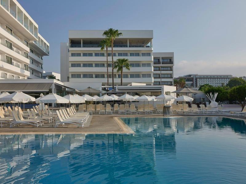 Moderner Hotelkomplex mit Pool, Sonnenliegen und Sonnenschirmen, umgeben von Palmen bei klarem Himmel.