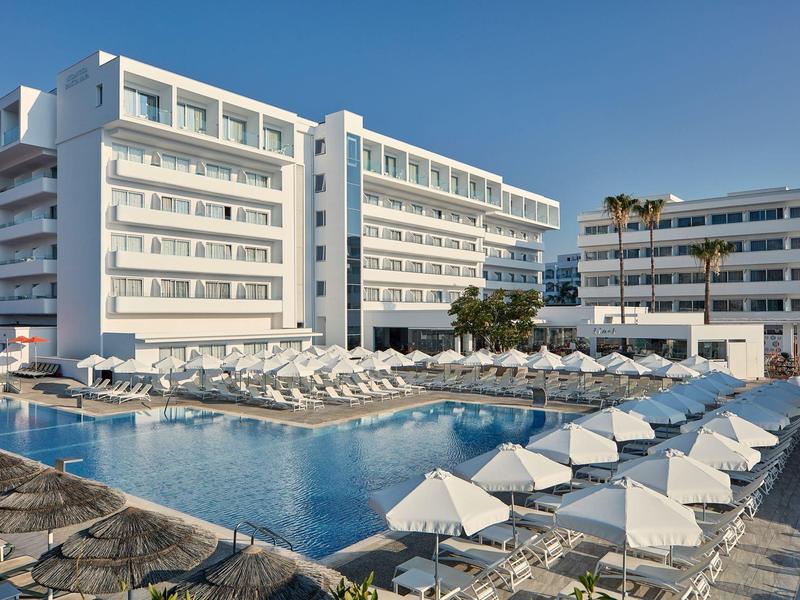 Großes modernes Hotel mit Pool, vielen Liegestühlen und Sonnenschirmen bei klarem Himmel.
