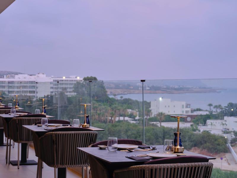Restaurant mit Tischen und Stühlen auf Terrasse, Blick auf Meer und Stadt bei bewölktem Himmel.
