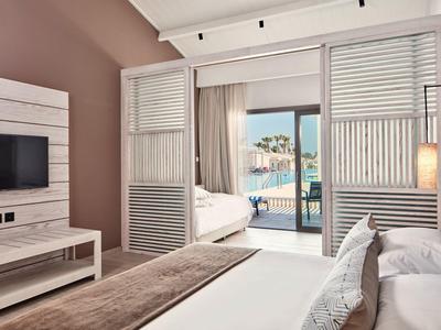 Schlafzimmer mit Doppelbett, braun-weißer Einrichtung, TV und Blick durch Schiebefenster auf Pool und Meer.