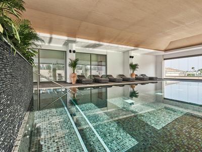 Moderner Innenpool mit Glaswand, grünem Mosaik, Holzdecke und Liegen am Rand.