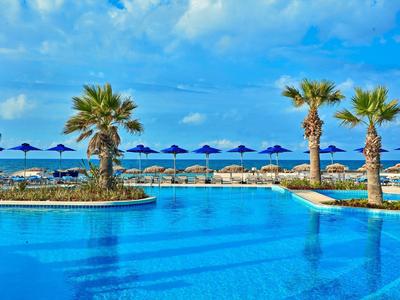 Großer blauer Pool mit Palmen und blauen Sonnenschirmen am Strand unter wolkigem Himmel.