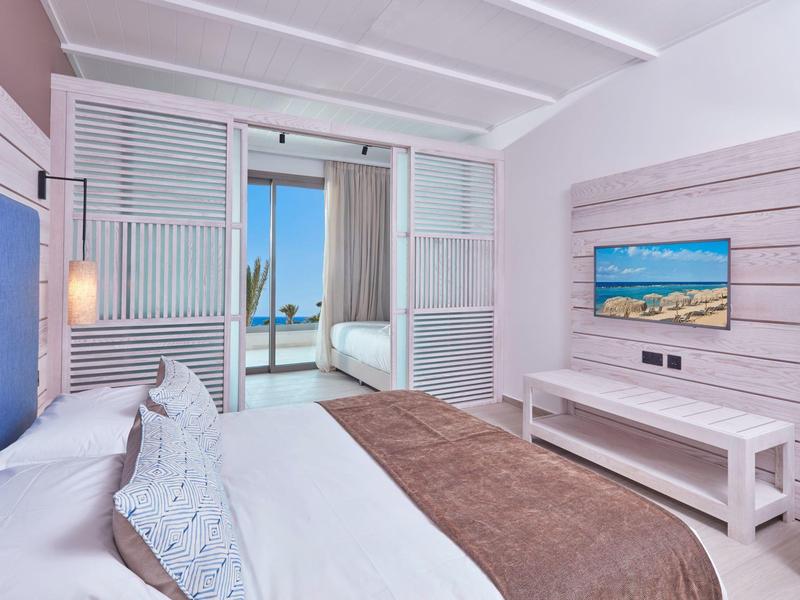 Helles Schlafzimmer mit Doppelbett, TV an der Wand und Blick durch große Fenstertür auf Meer.