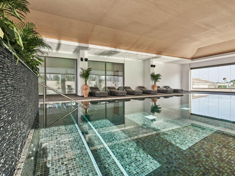 Moderner Innenpool mit Glaswand, grünem Mosaik, Holzdecke und Liegen am Rand.