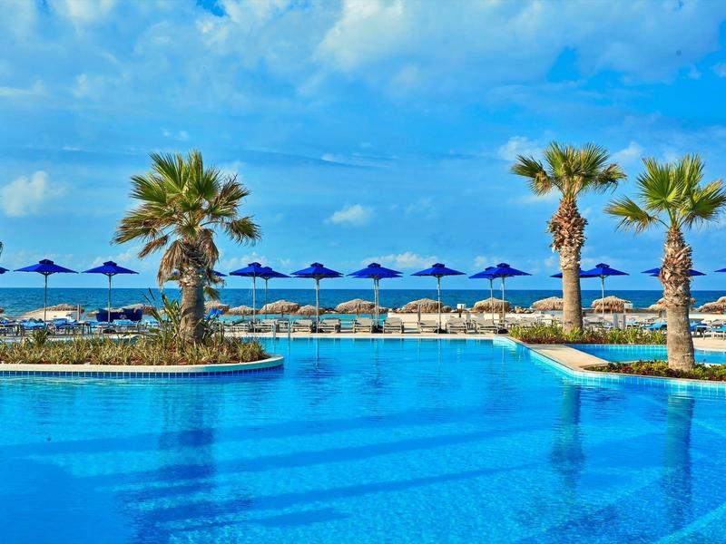 Großer blauer Pool mit Palmen und blauen Sonnenschirmen am Strand unter wolkigem Himmel.