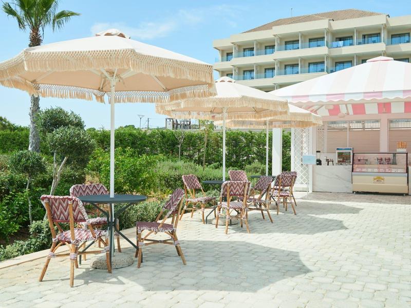 Terrasse mit runden Tischen, geflochtenen Stühlen und großen Sonnenschirmen vor Gebäude.