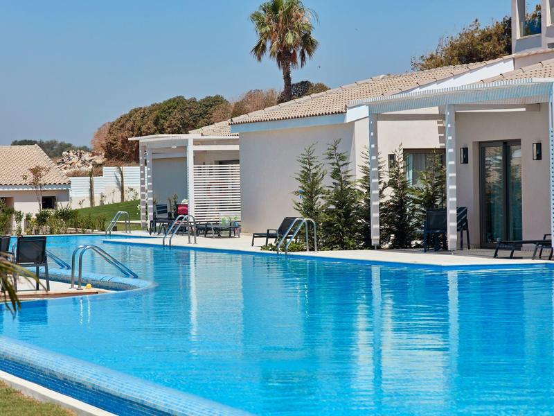 Großer blauer Pool vor weißen Bungalows mit Palmbaum und klar blauem Himmel.