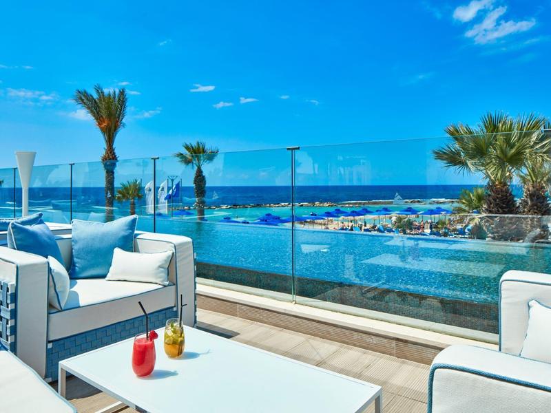 Moderne Terrasse mit weißen Sitzmöbeln, Glasgeländer und Meerblick bei blauem Himmel.