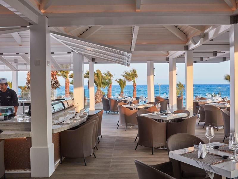 Offenes Restaurant am Meer mit gedeckten Tischen, Holzboden und weißen Säulen unter Dach.
