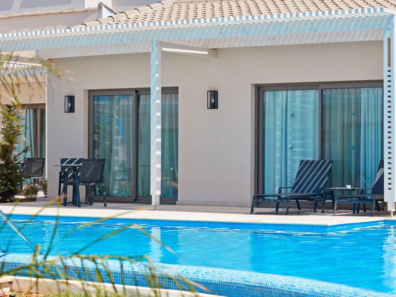 Moderner Bungalow mit Terrasse und Pool, blau-weiße Stühle und große Glas-Schiebetüren.