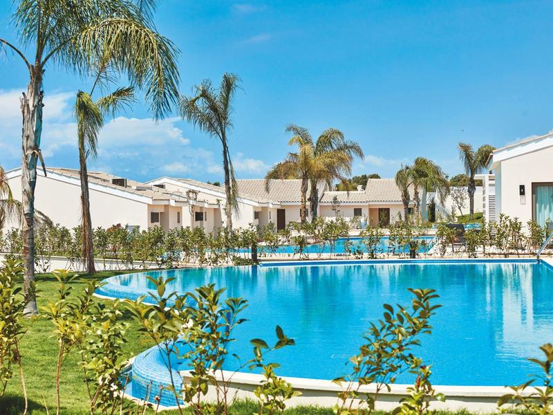 Großer Pool mit Palmen, blauer Himmel und weißen Gebäuden in einer gepflegten Anlage.