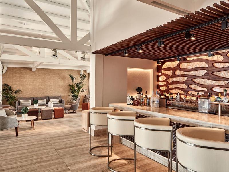 Moderner Barbereich mit Holztheke, weißen Barhockern und gemütlichen Sitzgruppen in hellem Ambiente.