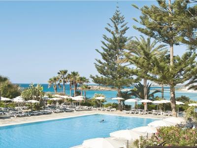 Großer blauer Pool mit Sonnenliegen, Palmen und Blick auf das Meer unter klarem Himmel.