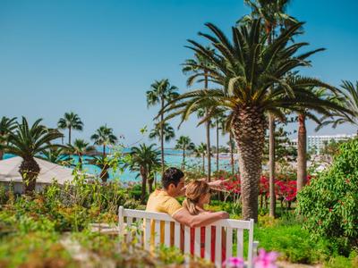 Couple assis sur un banc dans un jardin tropical avec des palmiers et un ciel bleu.
