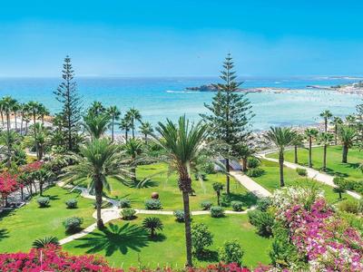 Blick auf einen grünen Park mit Palmen, blühenden Sträuchern und dahinter das blaue Meer bei sonnigem Himmel.
