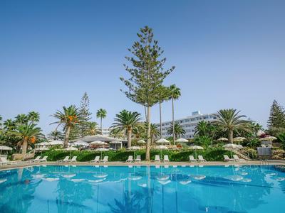 Vue sur une piscine d'hôtel avec des palmiers et des chaises longues sous un ciel dégagé.