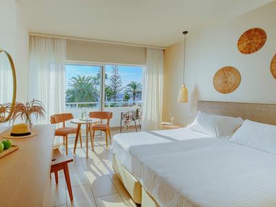 Modernes, helles Hotelzimmer mit zwei Betten, Holzmöbeln, Balkon und Blick auf Palmen bei Sonnenschein.