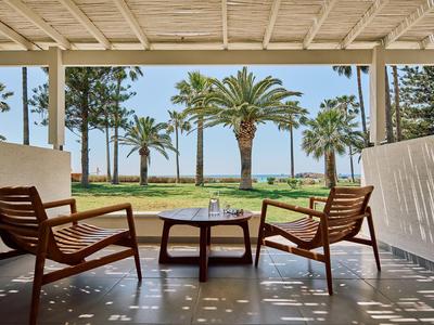 Zwei Holzstühle und Tisch auf Veranda mit Blick auf Palmen und Meer im Hintergrund.