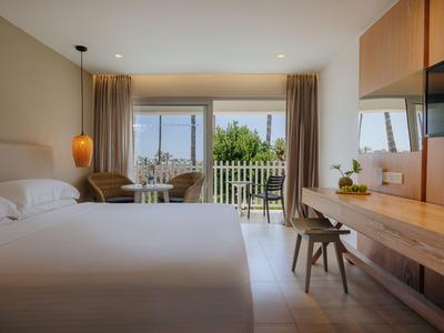 Helles Hotelzimmer mit großem Bett, Holzmöbeln und Blick auf einen Balkon mit grüner Landschaft.