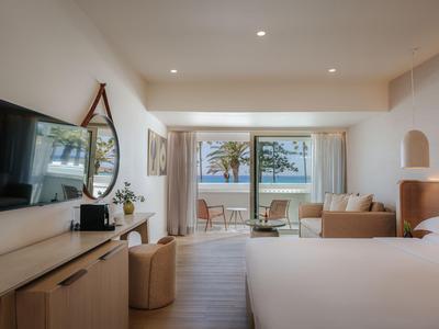 Helles Hotelzimmer mit Doppelbett, Sofa, TV, Spiegel und Balkon mit Meerblick.