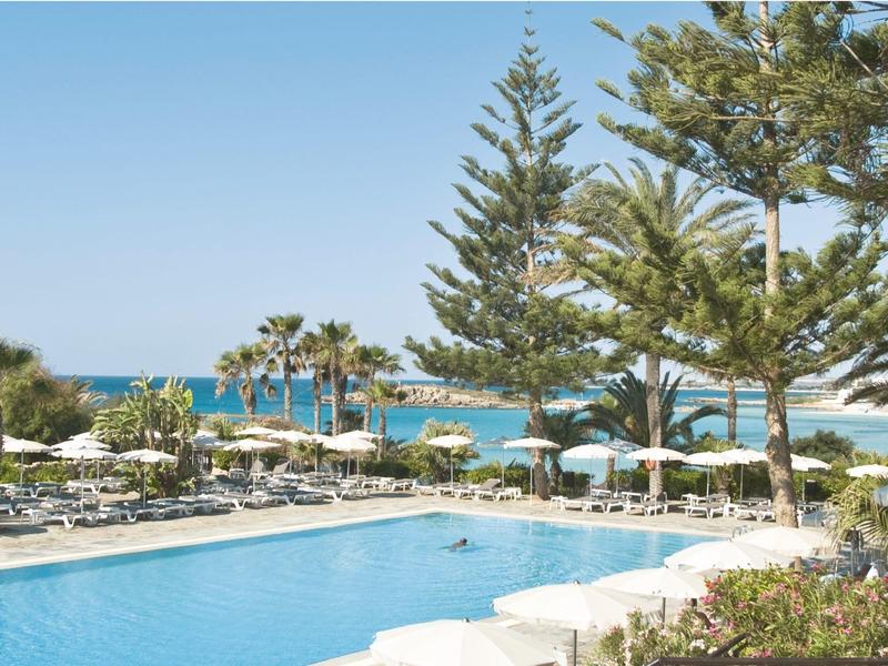 Großer blauer Pool mit Sonnenliegen, Palmen und Blick auf das Meer unter klarem Himmel.