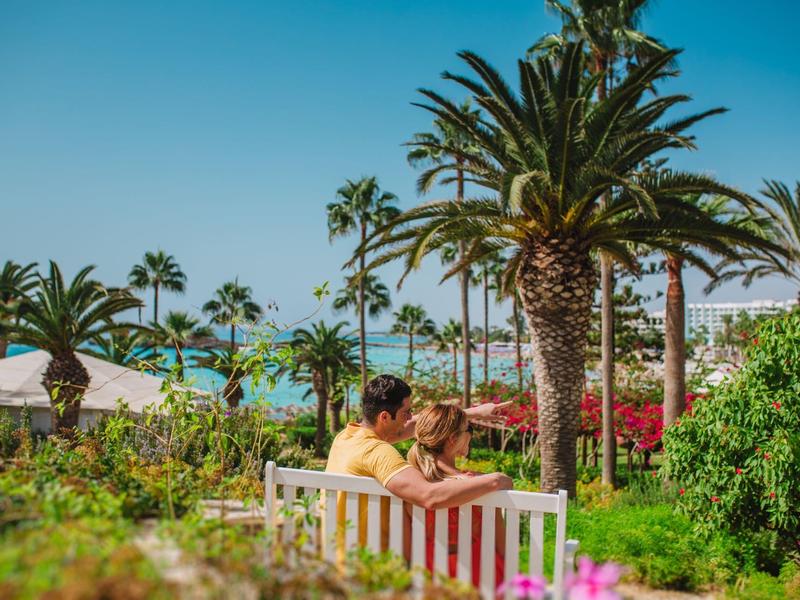 Zwei Personen sitzen auf einer Bank im Grünen, umgeben von Palmen unter blauem Himmel.
