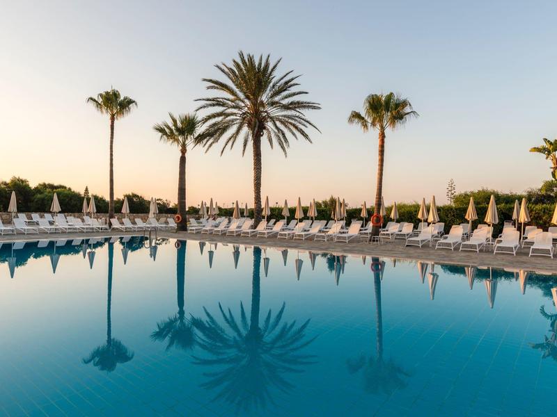 Großer, ruhiger Pool mit weißen Liegestühlen und Palmen bei Sonnenuntergang am klaren Himmel.
