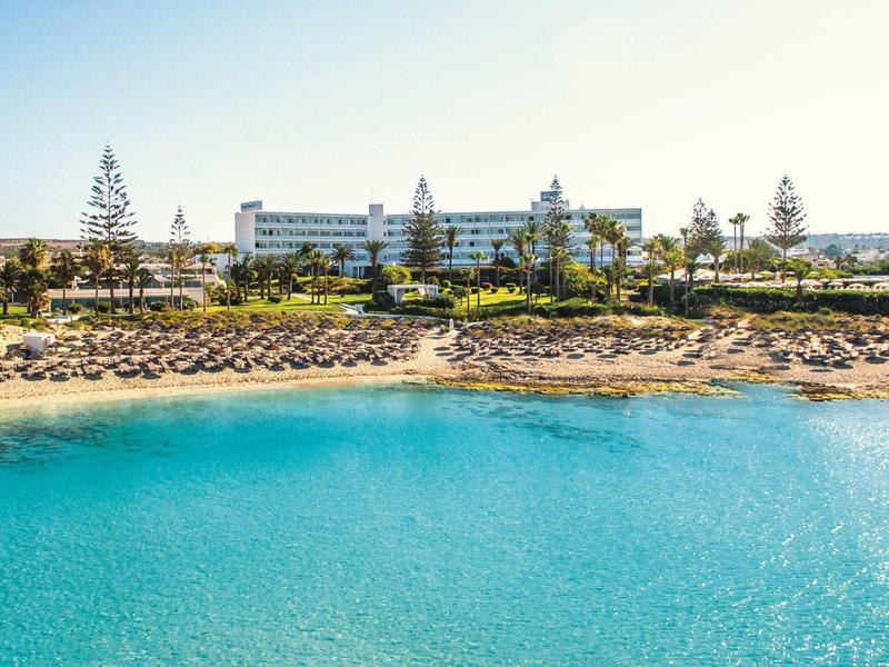 Klares türkisfarbenes Meer, Sandstrand mit Liegestühlen und Palmen vor großem Hotelgebäude am Horizont.