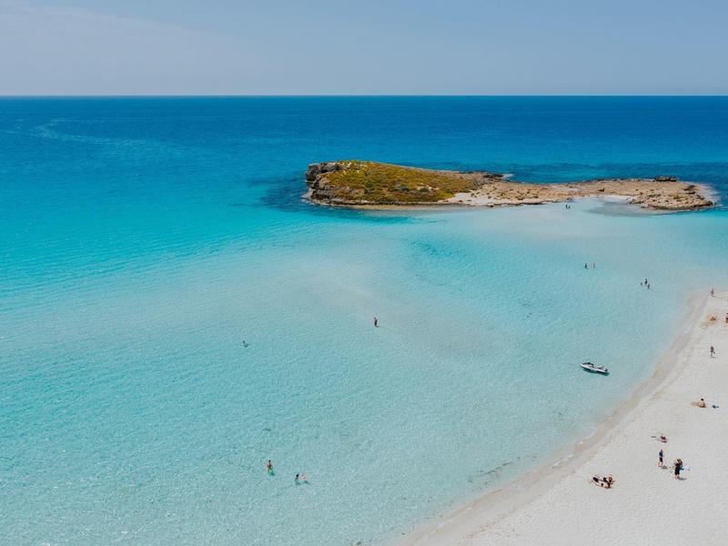Klares türkises Wasser und weißer Sandstrand mit kleiner Insel im Hintergrund unter blauem Himmel.
