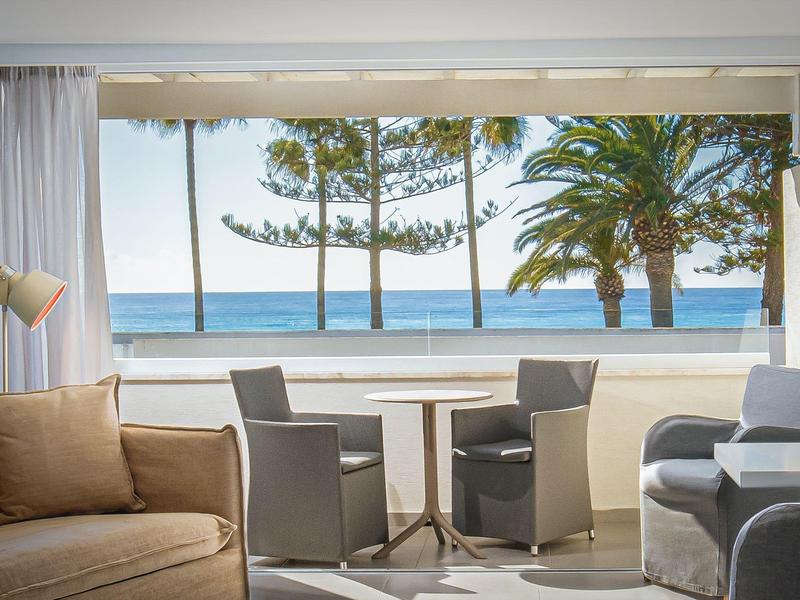 Wohnzimmer mit beiger Couch, grauen Stühlen, Tisch und großem Fenster mit Meerblick und Palmen.