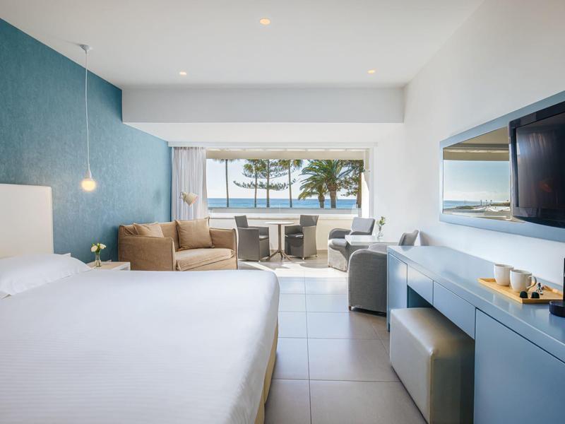 Helles Hotelzimmer mit großem Bett, Sitzbereich, Balkon und Meerblick. Weiße und blaue Farben dominieren.