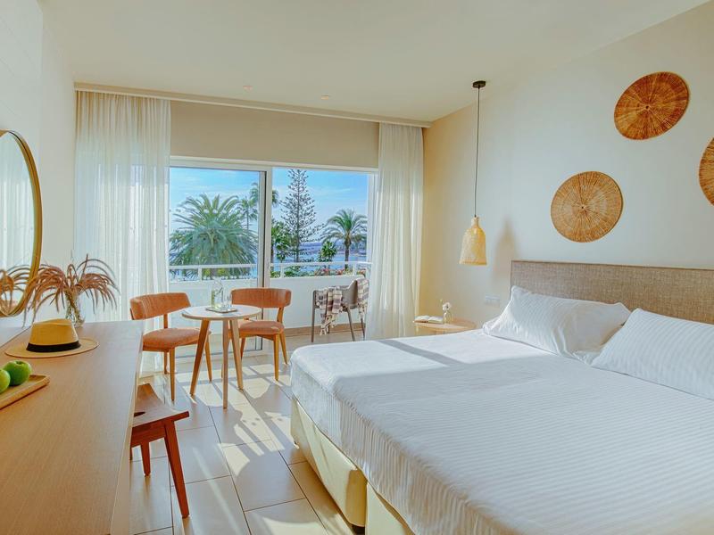 Modernes, helles Hotelzimmer mit zwei Betten, Holzmöbeln, Balkon und Blick auf Palmen bei Sonnenschein.