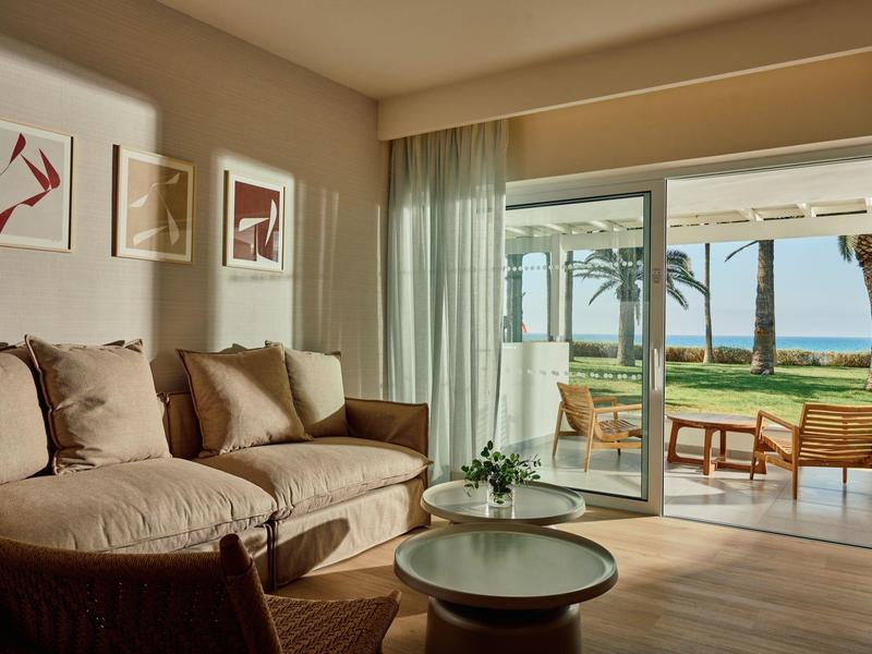 Helles Wohnzimmer mit gemütlichem Sofa, Holzstuhl, rundem Couchtisch und Blick auf Terrasse mit Meer.